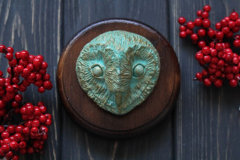 May include: A teal-colored owl face sculpture mounted on a round, dark wooden plaque. The owl has detailed feather textures and large, round eyes. Red berries are arranged on either side of the owl, against a dark wood background.