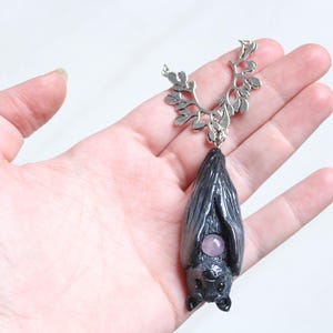 May include: A handcrafted bat pendant necklace. The pendant is dark gray with a small, round, purple gemstone. The necklace has a silver-colored, leaf-like design at the top. The bat pendant is approximately 2 inches long.