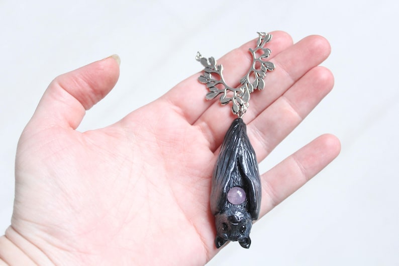 May include: A handcrafted bat pendant necklace. The pendant is dark gray with a small, round, purple gemstone. The necklace has a silver-colored, leaf-like design at the top. The bat pendant is approximately 2 inches long.