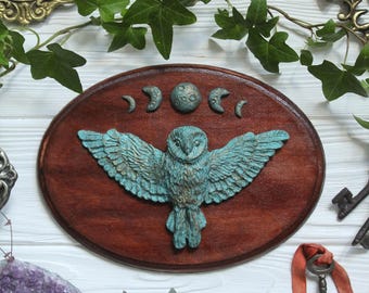 May include: An oval wooden plaque featuring a sculpted teal owl with outstretched wings positioned below a series of five moon phases. The owl has detailed feather textures and a weathered finish, mounted on a dark brown stained wood base.