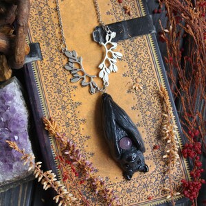 May include: A silver necklace with a bat pendant. The bat is black with a purple gemstone and hangs from a silver chain with a leaf design. The necklace rests on a vintage book with a floral pattern, surrounded by dried flowers.