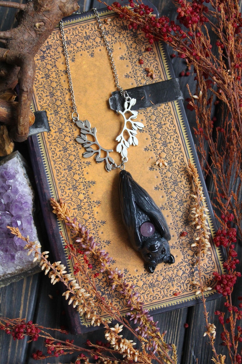 May include: A silver necklace with a bat pendant. The bat is black with a purple gemstone and hangs from a silver chain with a leaf design. The necklace rests on a vintage book with a floral pattern, surrounded by dried flowers.