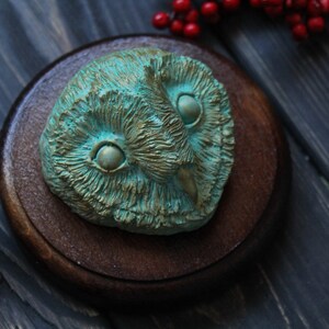 May include: A detailed, teal-colored owl sculpture with a textured surface, mounted on a round, dark brown wooden base. The owl's eyes are prominent, and the sculpture has a weathered, antique appearance. Red berries are in the background.