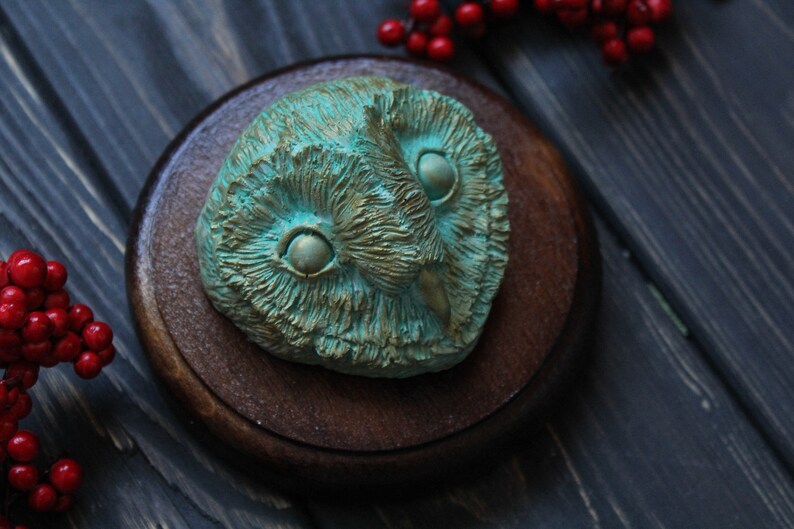 May include: A detailed, teal-colored owl sculpture with a textured surface, mounted on a round, dark brown wooden base. The owl's eyes are prominent, and the sculpture has a weathered, antique appearance. Red berries are in the background.