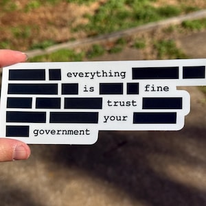 May include: A white sticker with black text that reads "everything is fine trust your government." The text is arranged in a block-style design with some words partially obscured by black rectangles.