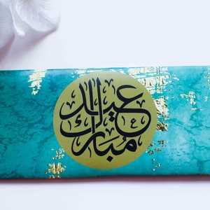 May include: A teal and gold envelope with a gold circle containing black Arabic calligraphy that reads "Eid Mubarak".