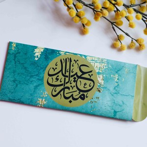 May include: A turquoise and gold envelope with a circular gold sticker featuring black Arabic calligraphy that reads "Eid Mubarak".