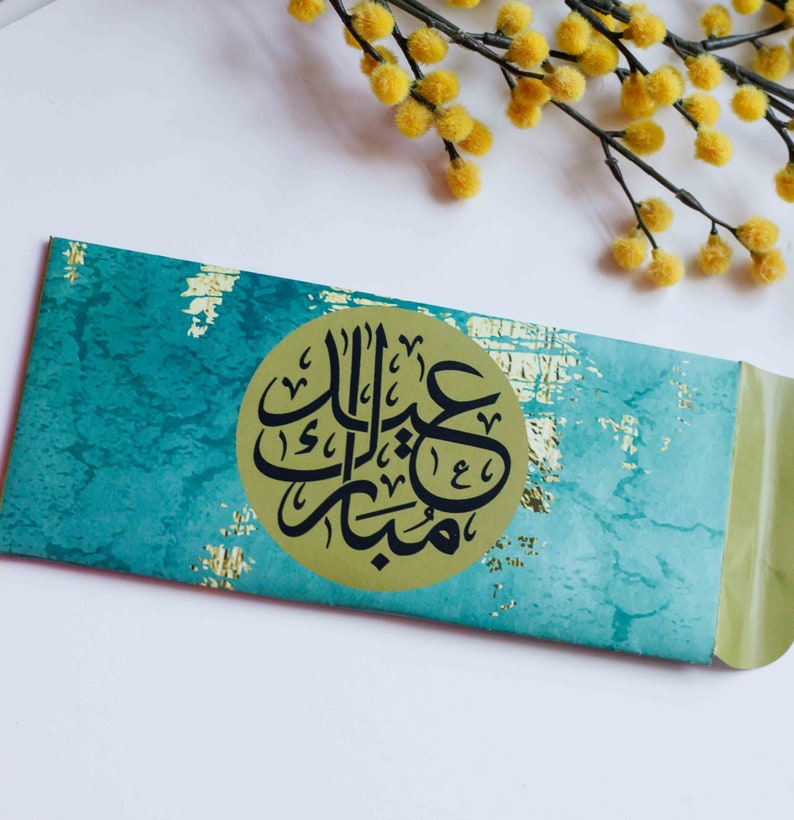 May include: A turquoise and gold envelope with a circular gold sticker featuring black Arabic calligraphy that reads "Eid Mubarak".