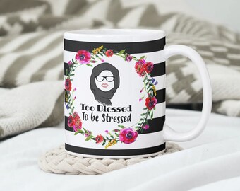 May include: White ceramic mug with black stripes and a floral wreath design. The wreath features pink, red, and yellow flowers. The mug has a black and white illustration of a woman wearing a headscarf and glasses. The text "Too Blessed To Be Stressed" is printed on the mug.