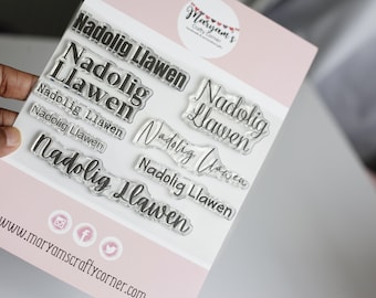 May include: Clear acrylic stamp set with the Welsh phrase "Nadolig Llawen" (Merry Christmas) in various fonts and sizes. The stamps are arranged on a white background with a pink border. The website address "www.maryamscraftycorner.com" is printed at the bottom.