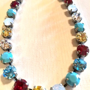 May include: A silver necklace with a rainbow of oval-shaped gemstones. The necklace features turquoise, blue, red, yellow, and white stones.