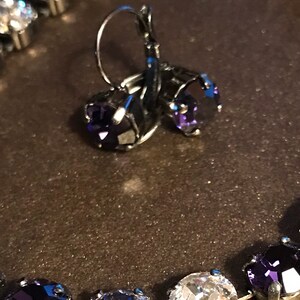 May include: A pair of silver earrings with dangling purple crystal stones. The earrings are shown with a silver necklace with alternating clear and purple crystal stones.