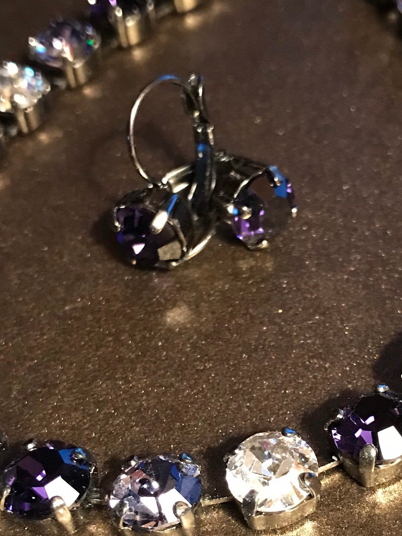 May include: A pair of silver earrings with dangling purple crystal stones. The earrings are shown with a silver necklace with alternating clear and purple crystal stones.