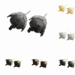 May include: A set of six pairs of earring settings in various finishes, including black, silver, gold, and antique bronze. Each setting features a round bezel with a pronged edge, designed to hold a round stone or cabochon.