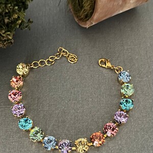 Puede incluir: Un brazalete de cadena dorada con un arcoíris de cristales redondos y facetados. Los cristales están dispuestos en un patrón graduado, con el cristal más grande en la parte superior y el cristal más pequeño en la parte inferior.