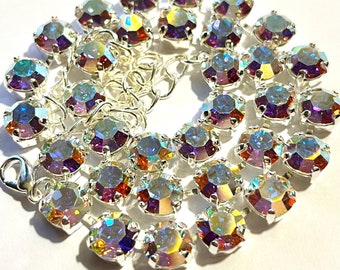May include: A silver-toned necklace featuring iridescent, round-cut crystals. The crystals have a rainbow effect, reflecting colors like purple, blue, and yellow. The necklace is secured with a lobster clasp.