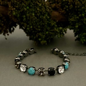 Puede incluir: Una pulsera de plata con una fila de cristales coloridos. La pulsera tiene un cristal turquesa, gris, negro y transparente.