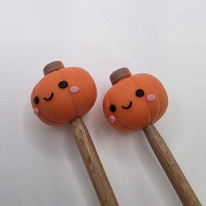 May include: Two orange pumpkin-shaped toppers with smiling faces and brown stems on wooden sticks. The pumpkins have pink cheeks and black eyes. The background is plain white.
