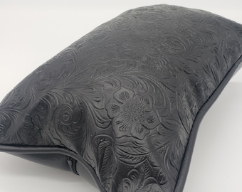 May include: Black leather cushion with an embossed floral pattern. The cushion has a zipper closure and a slightly curved shape.