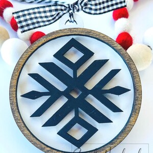 May include: A wooden round ornament with a black and white geometric snowflake design. The ornament is attached to a black and white checkered ribbon.