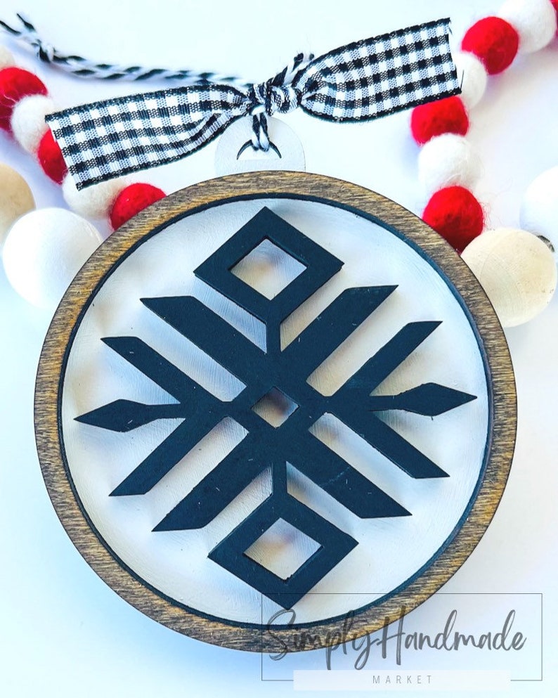 May include: A wooden round ornament with a black and white geometric snowflake design. The ornament is attached to a black and white checkered ribbon.