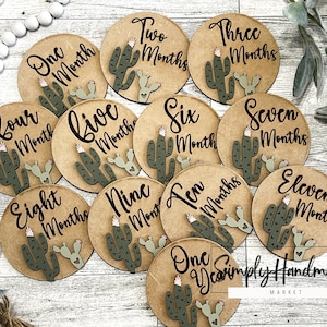May include: Twelve round wooden discs with cactus illustrations and the words "One Month" through "Eleven Months" and "One Year". The discs are arranged in a circle on a wooden surface.