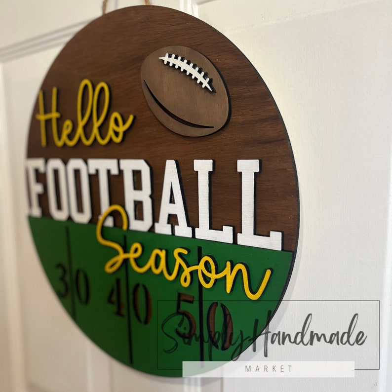 K&ouml;nnte beinhalten: Ein rundes Holzschild mit braunem Holzgrund und einem gr&uuml;nen und wei&szlig;en Fu&szlig;ballfeld-Design. Das Schild sagt "Hello Football Season" mit den Zahlen 30, 40 und 50 auf dem Feld. Das Schild hat auch einen braunen Fu&szlig;ballausschnitt.