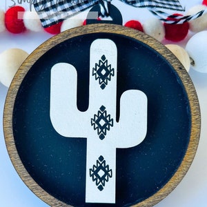 May include: A round wooden ornament with a black background and a white cactus design. The cactus has geometric patterns in black on it.