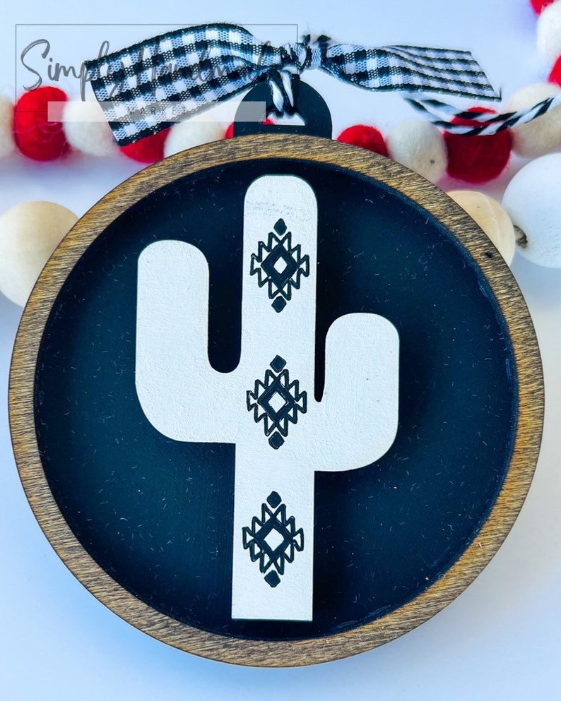 May include: A round wooden ornament with a black background and a white cactus design. The cactus has geometric patterns in black on it.