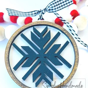 May include: A wooden round ornament with a dark teal snowflake design inside. The ornament is surrounded by a brown wooden frame. The ornament is hanging from a black and white checkered ribbon.