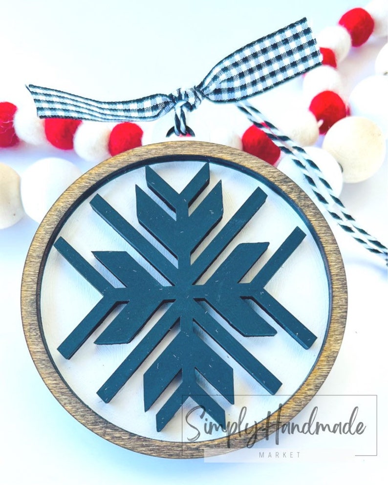 May include: A wooden round ornament with a dark teal snowflake design inside. The ornament is surrounded by a brown wooden frame. The ornament is hanging from a black and white checkered ribbon.