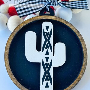 May include: A wooden round ornament with a black background and a white cactus with a geometric pattern. The ornament is attached to a black and white checkered ribbon.