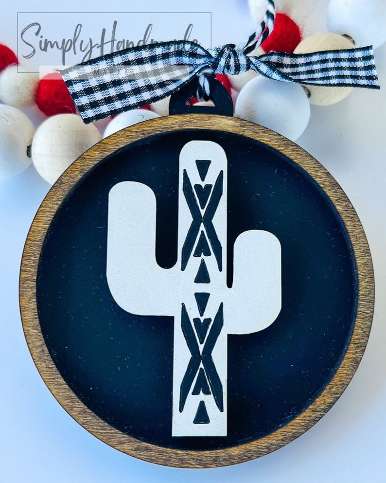 May include: A wooden round ornament with a black background and a white cactus with a geometric pattern. The ornament is attached to a black and white checkered ribbon.
