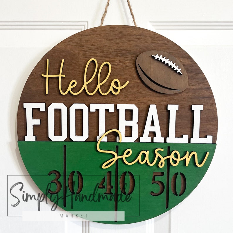 K&ouml;nnte beinhalten: Ein rundes Holzschild mit dem Text "Hello Football Season" in gelben und wei&szlig;en Buchstaben. Das Schild ist mit einem braunen Fu&szlig;ball und gr&uuml;n-wei&szlig;en Streifen verziert. Die Zahlen 30, 40 und 50 sind ebenfalls auf dem Schild zu sehen.