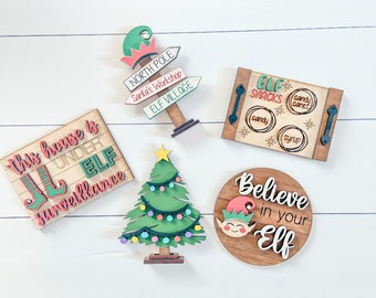 May include: Five wooden Christmas signs with a festive elf theme. The signs feature phrases like "This house is under elf surveillance", "Elf Snacks", "North Pole, Santa's Workshop, Elf Village", and "Believe in your Elf".