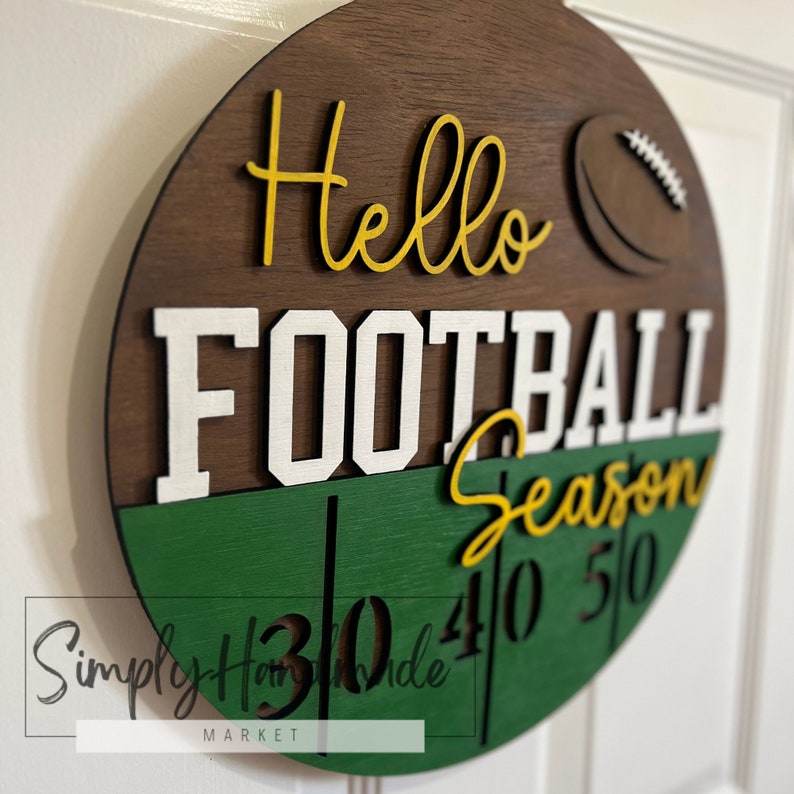 K&ouml;nnte beinhalten: Ein rundes Holzschild mit den Worten "Hello Football Season" in gelben und wei&szlig;en Buchstaben. Das Schild ist mit einem braunen Fu&szlig;ball und gr&uuml;n-wei&szlig;en Streifen verziert. Die Zahlen "30", "40" und "50" sind auf dem Schild sichtbar.