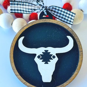 May include: A wooden ornament with a black background and a white bull's head silhouette. The bull's head has a geometric pattern on its forehead. The ornament is decorated with a black and white checkered ribbon.