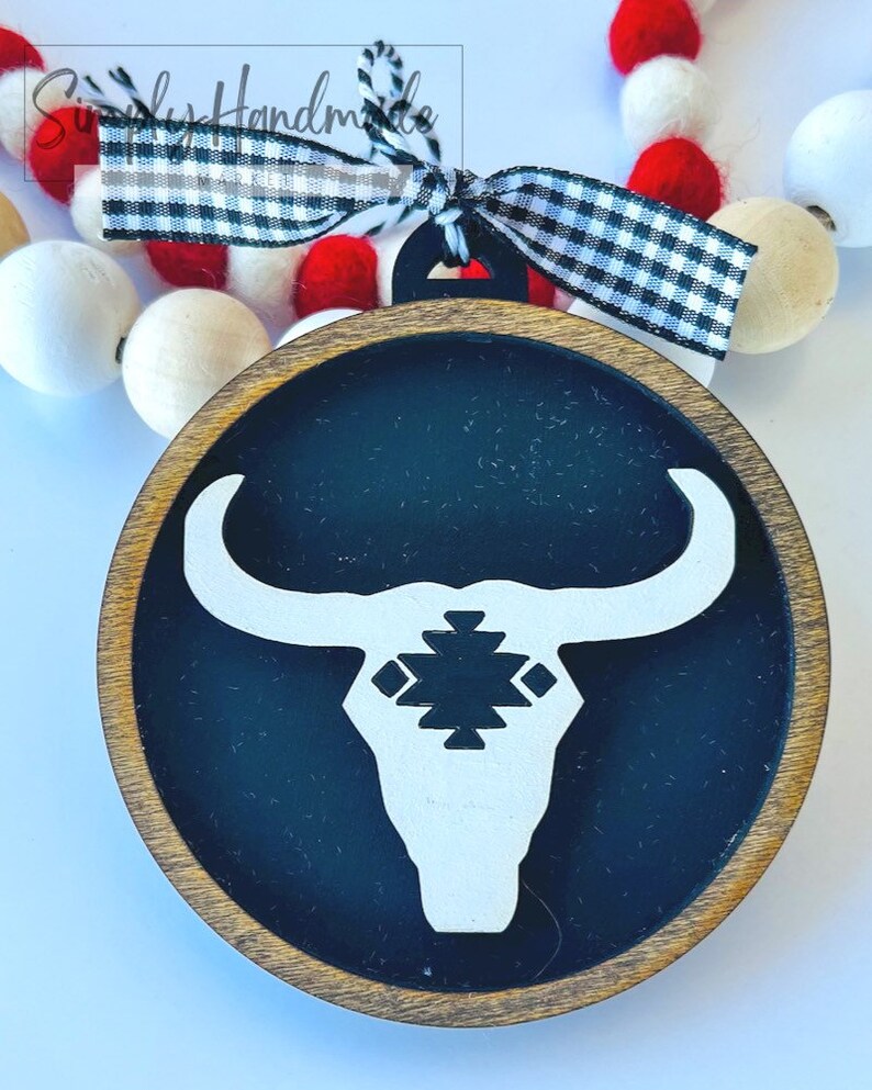 May include: A wooden ornament with a black background and a white bull's head silhouette. The bull's head has a geometric pattern on its forehead. The ornament is decorated with a black and white checkered ribbon.