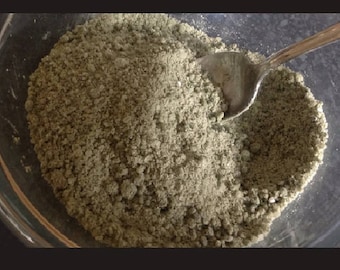 May include: A clear glass bowl filled with a light green, granular substance. A silver spoon rests in the substance, partially submerged. The bowl is set against a dark background.