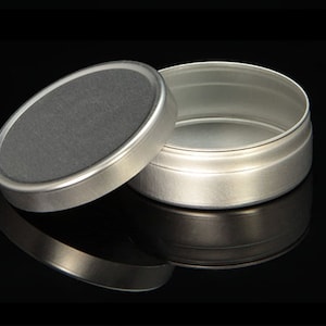 May include: A silver metal tin with a lid. The tin is round and has a smooth surface. The lid is slightly domed and has a smooth surface.