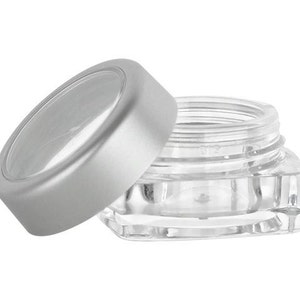 May include: A clear square plastic container with a silver lid. The lid has a clear plastic window on top.
