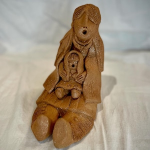 May include: A terracotta sculpture of a seated figure holding a smaller figure. The larger figure has long hair and is wearing a shawl. The smaller figure is also wearing a shawl. The sculpture is a warm brown color.