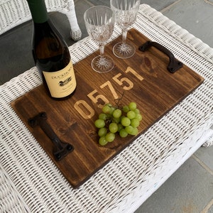 May include: A wooden serving tray with a brown stain and two metal handles. The tray has the number "05751" engraved on it. There is a bottle of white wine, a bunch of green grapes, and two clear wine glasses on the tray.