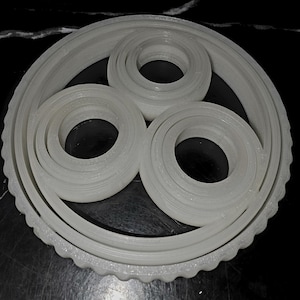 May include: A white, 3D-printed gear assembly with three circular components and a larger, toothed outer ring. The design features concentric circles and a clean, modern aesthetic. The gear is likely for a mechanical or engineering project.