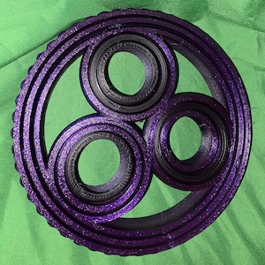 May include: A purple, circular, 3D-printed object with three central circular openings and a textured outer ring. The object has a glittery finish and is set against a green background.