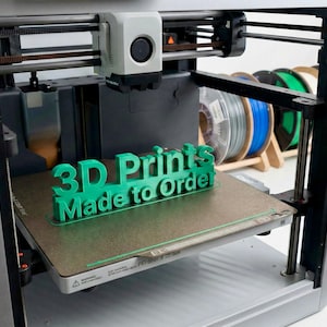 May include: A 3D printer in operation, with a green plastic sign that reads "3D Prints Made to Order" on the print bed. The printer is silver and black, with spools of filament in the background.