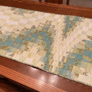 Könnte beinhalten: Ein Tischläufer mit Patchwork-Muster aus weißen, grünen und blauen Stoffen. Der Läufer hat einen gewellten Rand und ist aus einem weichen, gesteppten Material gefertigt.