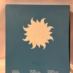 Op de afbeelding: Een blauwe boekomslag met een wit zonnesterontwerp. De tekst luidt "Star Atlas of Reference Stars and Nonstellar Objects" en "Smithsonian Astrophysical Observatory".