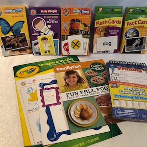May include: A collection of educational toys and books for children, including flash cards, a dry erase learning book, a coloring book, and a cookbook. The flash cards feature images of insects, people, traffic signs, dogs, and space. The dry erase learning book has a colorful cover with the words "Printing & Numbers". The coloring book has a green cover with the words "Squiggles & Doodles". The cookbook has a green cover with the words "Cooking with Kids" and a picture of fall food.