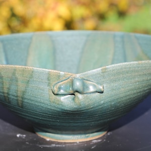 May include: A handcrafted teal ceramic bowl with a unique shape and two small handles. The bowl has a textured surface and a wide, open design. The teal color has subtle variations, adding depth to the piece. This decorative bowl is suitable for serving or display.